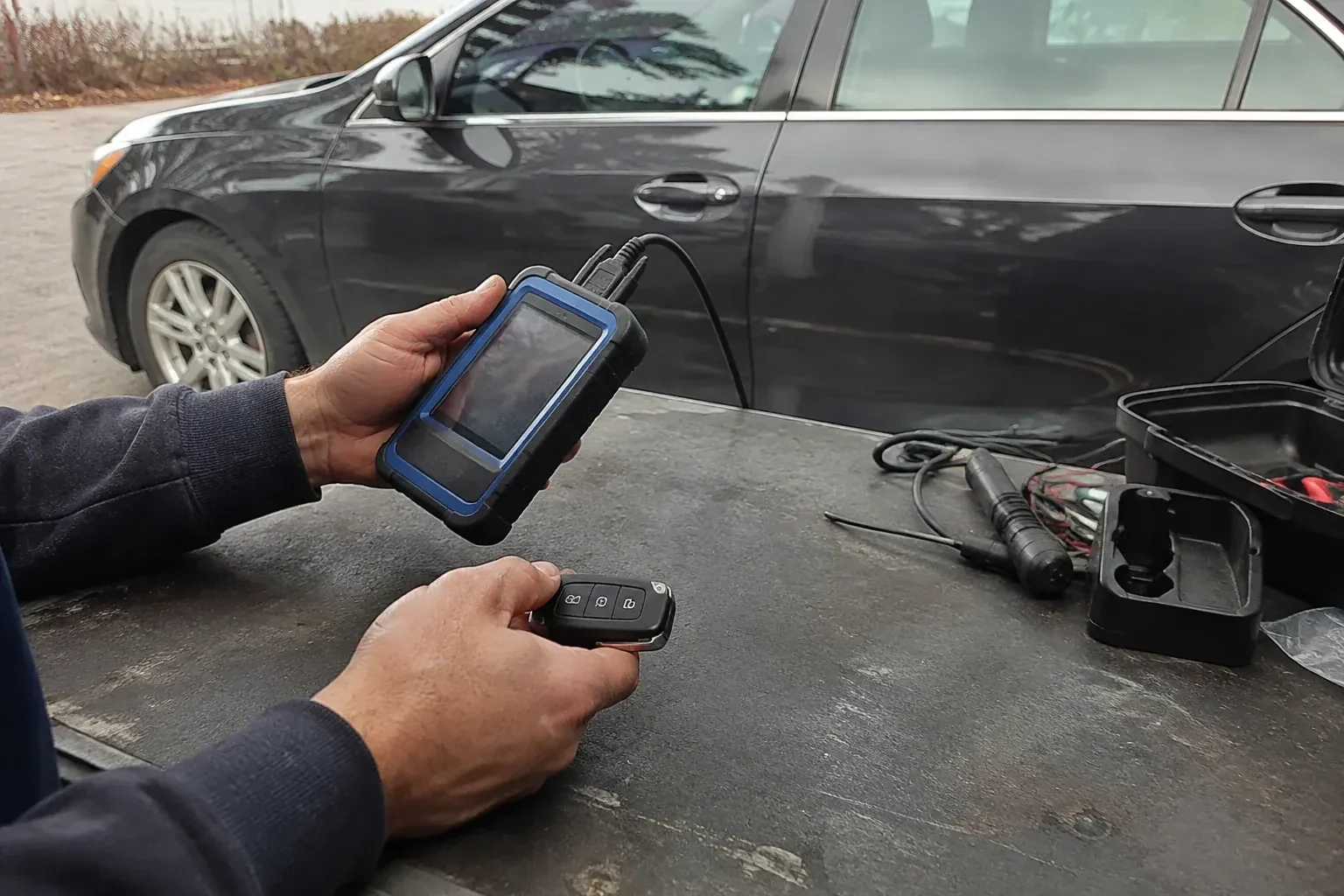 Hands holding a blue-edged electronic device and car-key fob at a worktable beside a parked car. — Transponder Key Programming, Peterborough