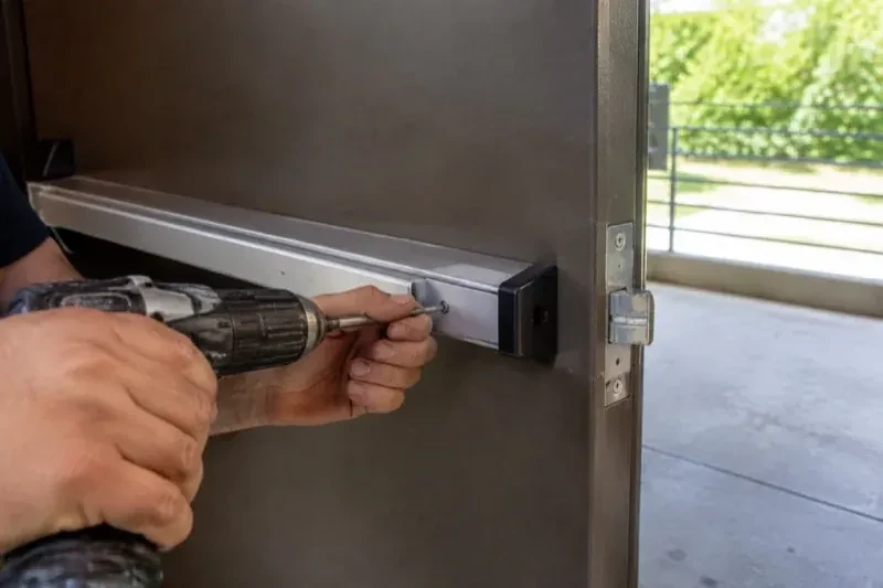 Hands using a power driver at the end of a horizontal push bar on an open metal door. — Panic & Exit Hardware, Peterborough